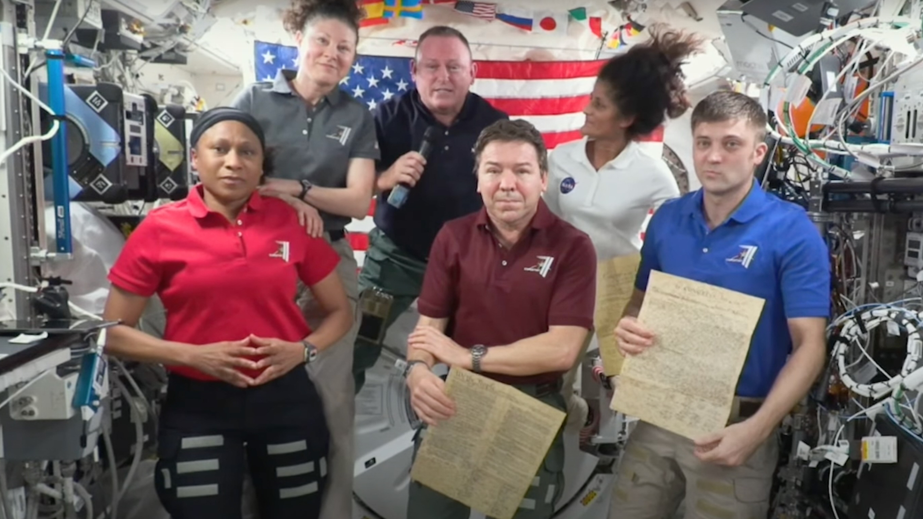 Four men and two women float aboard the International Space Station, with an American flag in the background