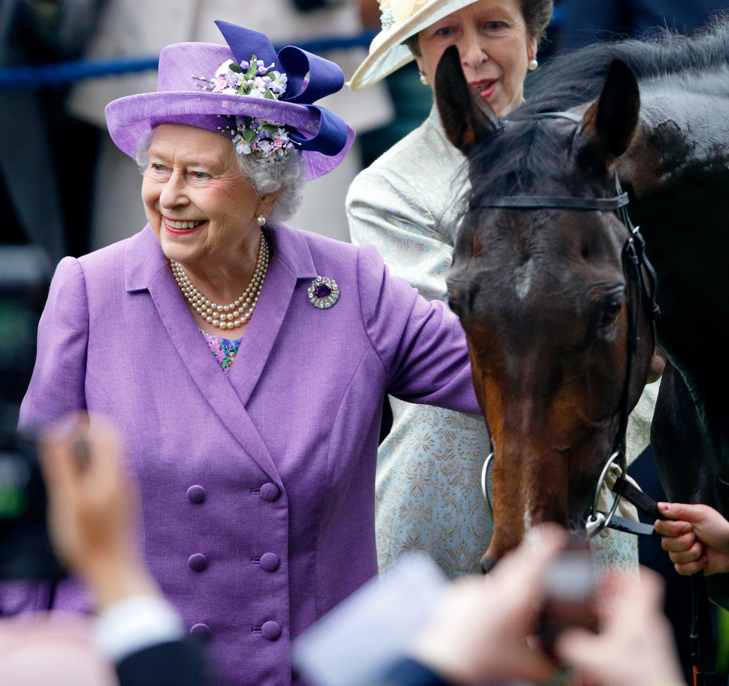 Queen Elizabeth wearing a purple suit smiling next to a horse