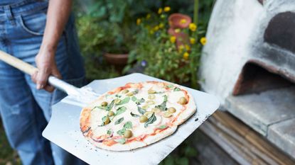 a woman taking out a pizza from a homemade pizza oven - GettyImages-1043643456