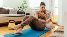 Woman exercising with kettlebell in domestic setting. She sits on an exercise mat with her feet raised and a kettlebell held to one side by her hip