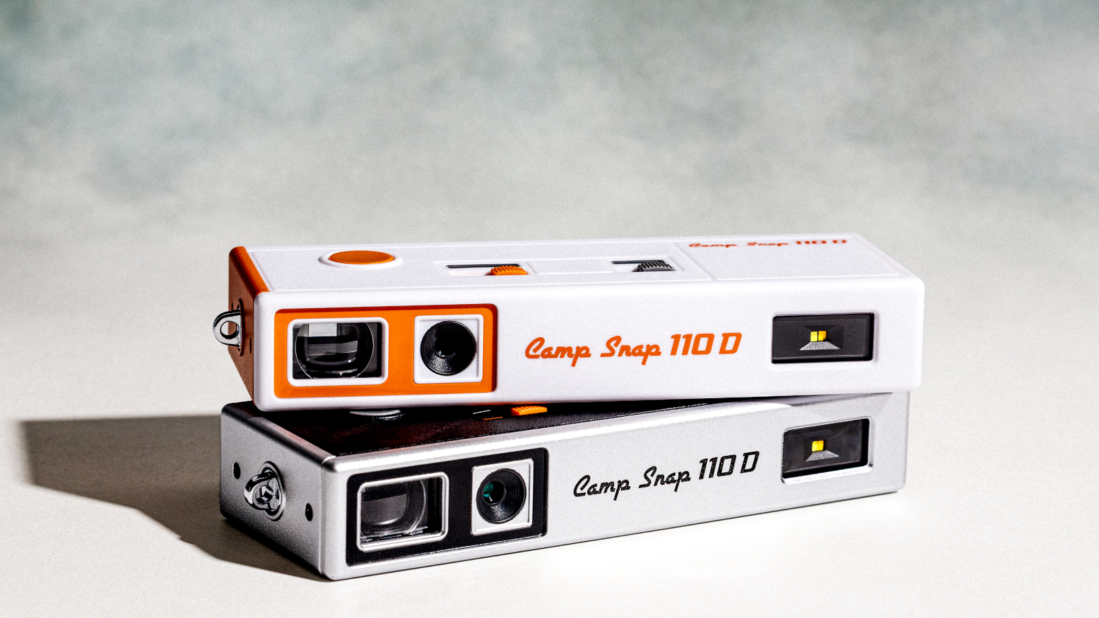Photograph of Camp Snap 110D digital camera in two colorways, white and black