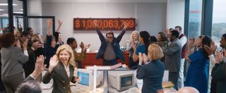 Ty (Zach Galifianakis) stands in front of an LED screen displaying a total of over one billion dollars. He has his arms raised triumphantly and is surrounded by Ty Co employees who are all applauding and celebrating.