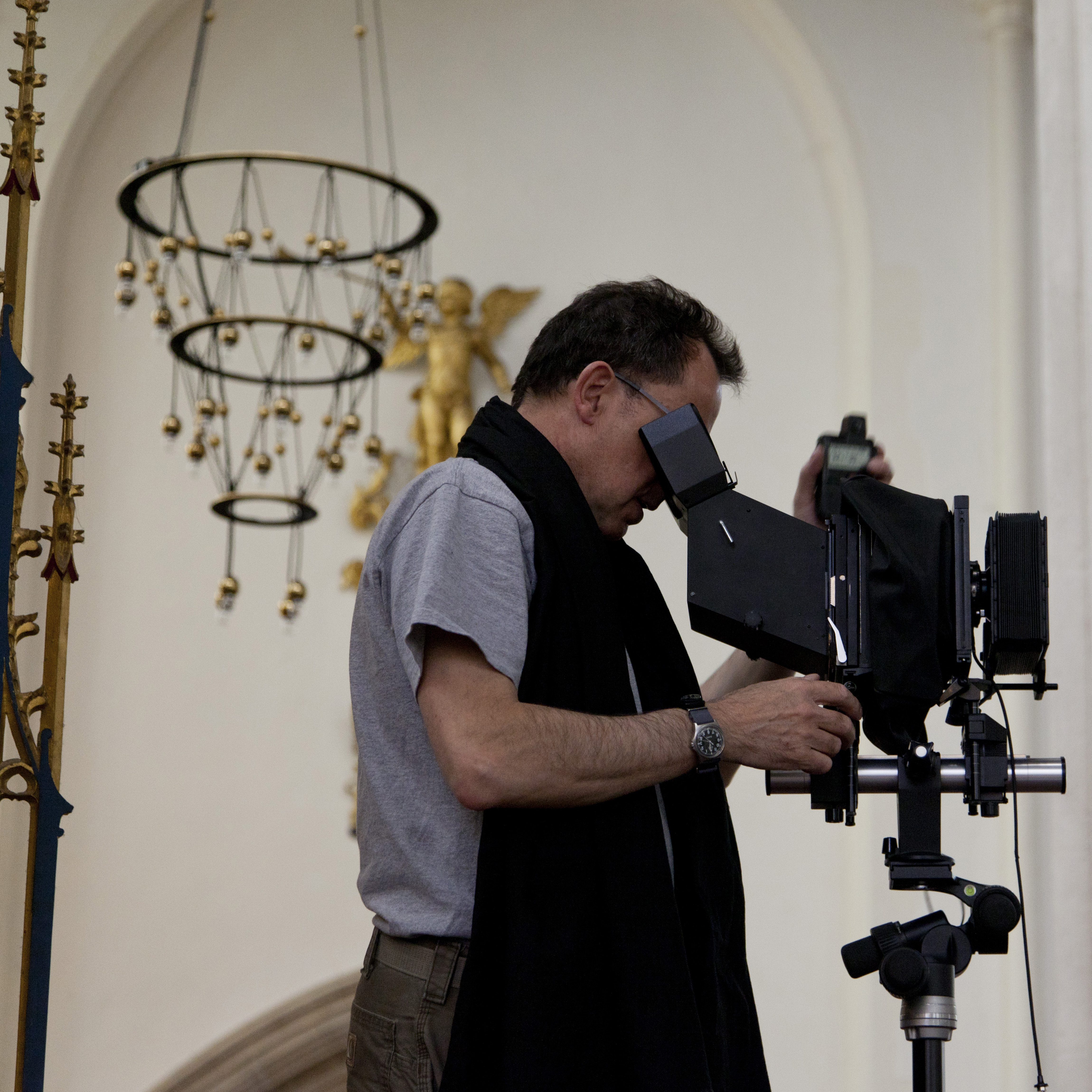 Peter Marlow looks through the viewfinder of a large format camera on a tripod inside St Edmundsbury Cathedral, beneath a gilded angel statue and hanging chandelier.