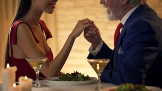 Younger woman in red dress holds takes hand of well dressed older man at dinner table.