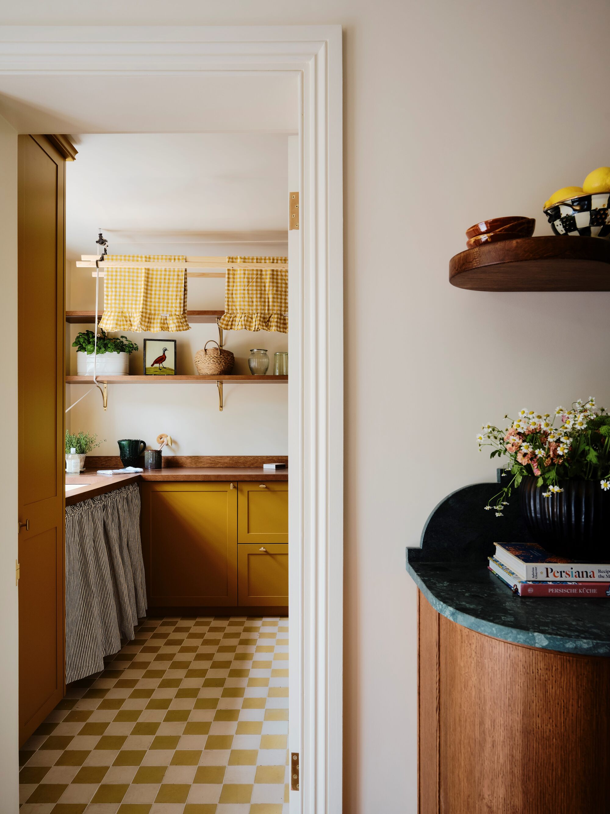 image of a yellow kitchen viewed from the next room. THe kitchen cabinets are painted yellow and the floors are a yellow and white checkerboard pattern. In the front room there is a wooden cabinet that has an emerald green marble top.