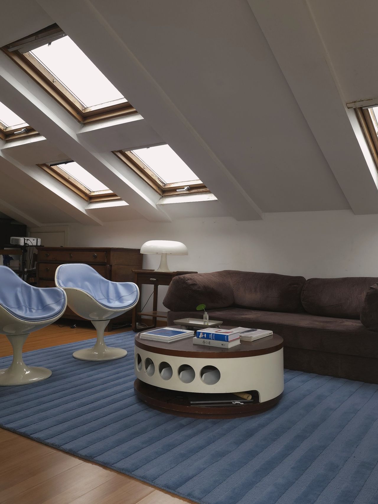 A mid-century modern-style living room with white slanted ceilings with skylights in them. There is a bug rug, a dark brown sofa, two blue and cream egg chairs, and a white wooden coffee table.