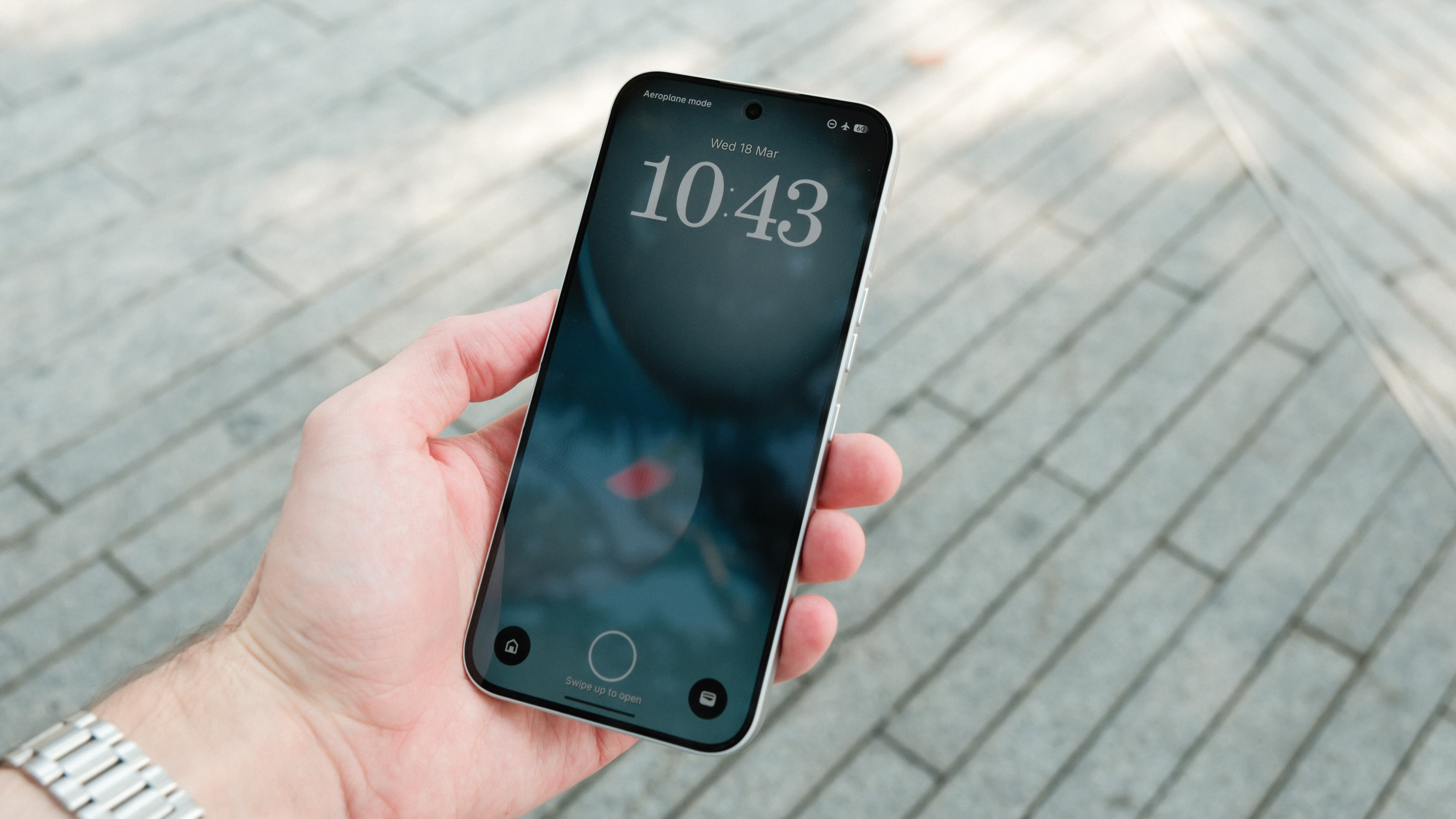 A person's hand holding a smartphone with a slim-bezel display. The lock screen shows a large digital clock reading 10:43 on a dark, moody wallpaper, with a &amp;quot;Swipe up to open&amp;quot; prompt at the bottom.