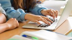 Female students in a classroom typing on a laptop keyboard with notepads in foreground.
