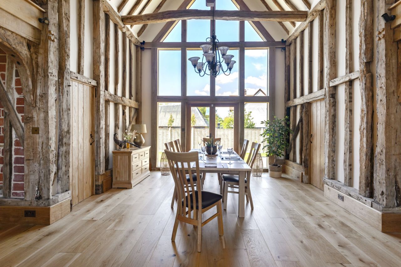 The interior of a modern barn conversion with exposed beams, a dining table and a two storey window