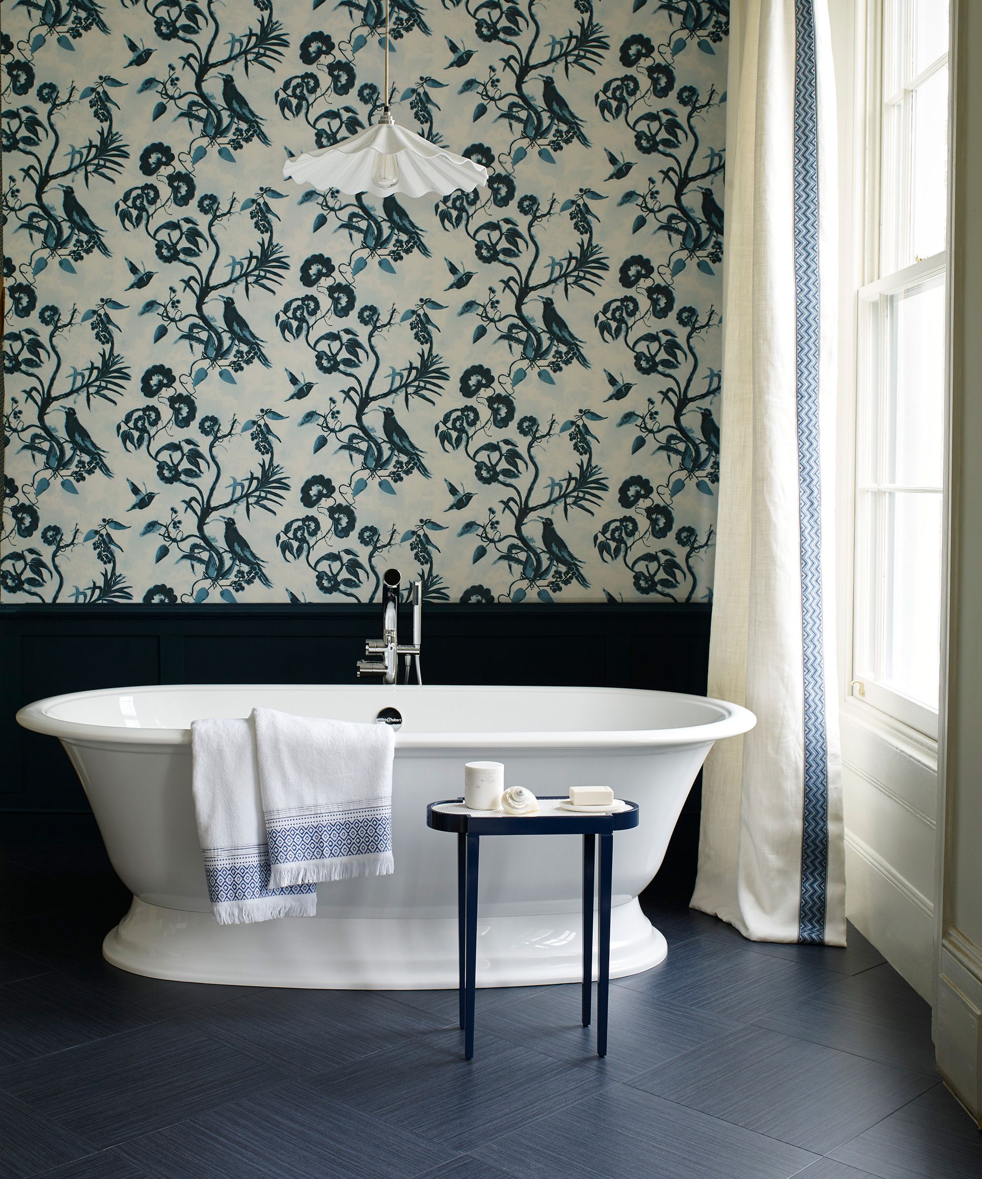 A freestanding white bath in a bathroom with floral blue and white wallpaper