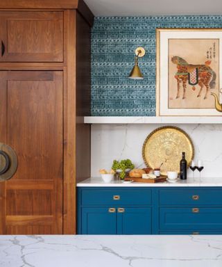 A kitchen with a tall dark brown wooden cabinet, a sink area with blue cabinets and a blue wall above it, and a marble kitchen island in front of it