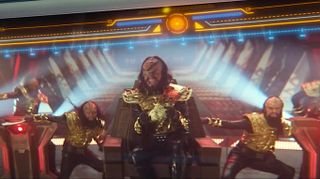 Five people in black and gold Klingon alien costumes dance and sing.