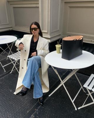 Sasha Mei wearing oversized sunglasses, a cream long coat, black sweater, blue jeans, tights, and black heels.