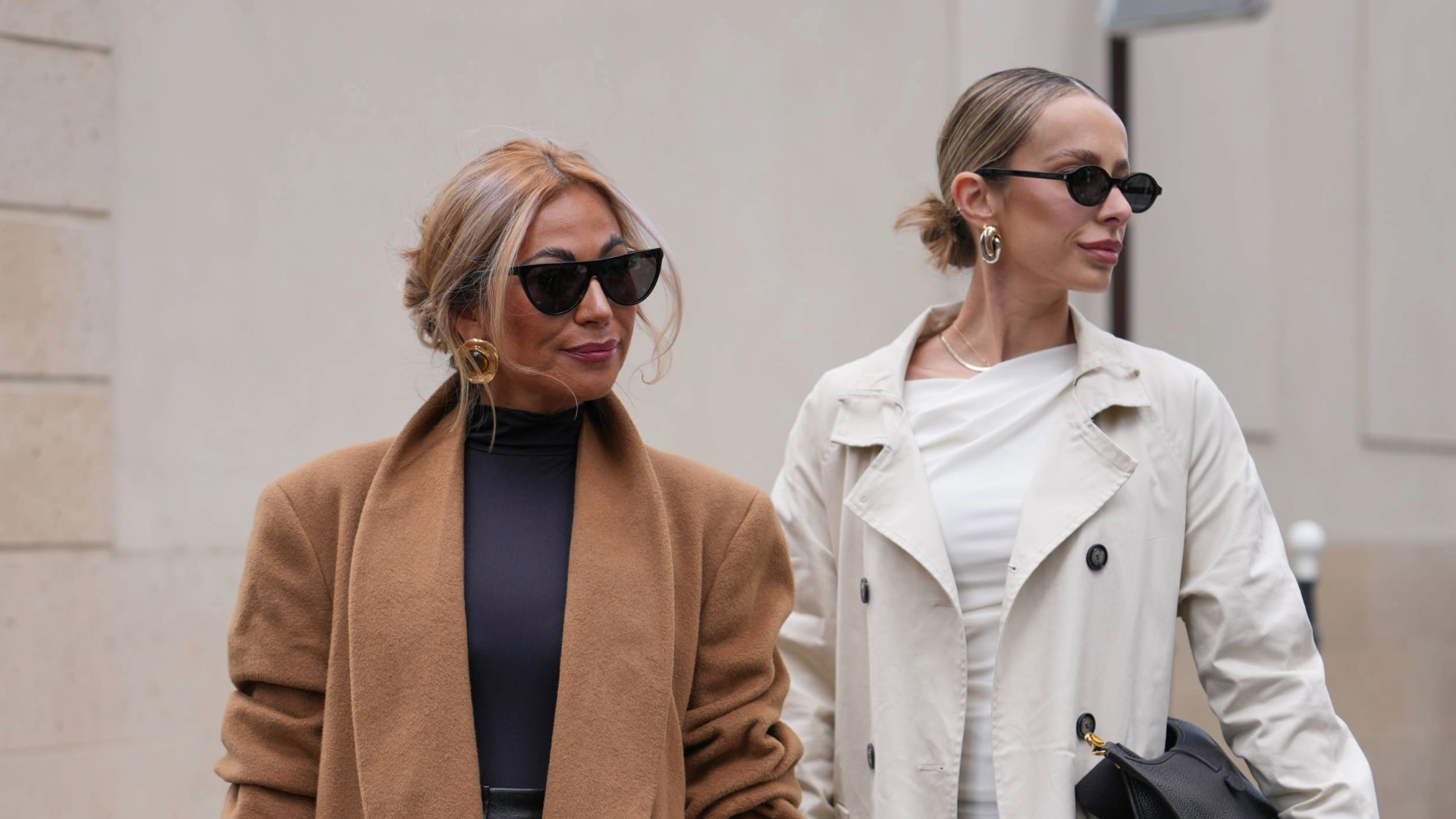 PARIS, FRANCE - OCTOBER 04: A guest (L) wears a loose low bun hairstyle with honey-blonde highlights and face-framing tendrils, black cat-eye sunglasses, large gold hoop earrings, a black leather clutch bag, a camel wool long double-breasted coat with wide lapels and black buttons over a black high-neck knit top and black leather trousers, brown suede gloves / mittens, black patent leather pointed-toe pumps shoes, A guest (R) wears a slicked-back low bun hairstyle with medium-length light brown hair and a center part, black oval sunglasses, chunky gold hoop earrings and a thin gold chain necklace, a black pebbled leather shoulder oversized Evelyne handbag from Hermes with gold hardware, a long beige cotton trench coat with double-breasted black buttons worn open over a white draped asymmetric top and black wide-leg trousers, black patent leather pointed-toe pumps shoes with stiletto heels, outside Hermes, during Paris Fashion Week - Womenswear Spring Summer 2026, on October 04, 2025 in Paris, France (Photo by Edward Berthelot/Getty Images)
