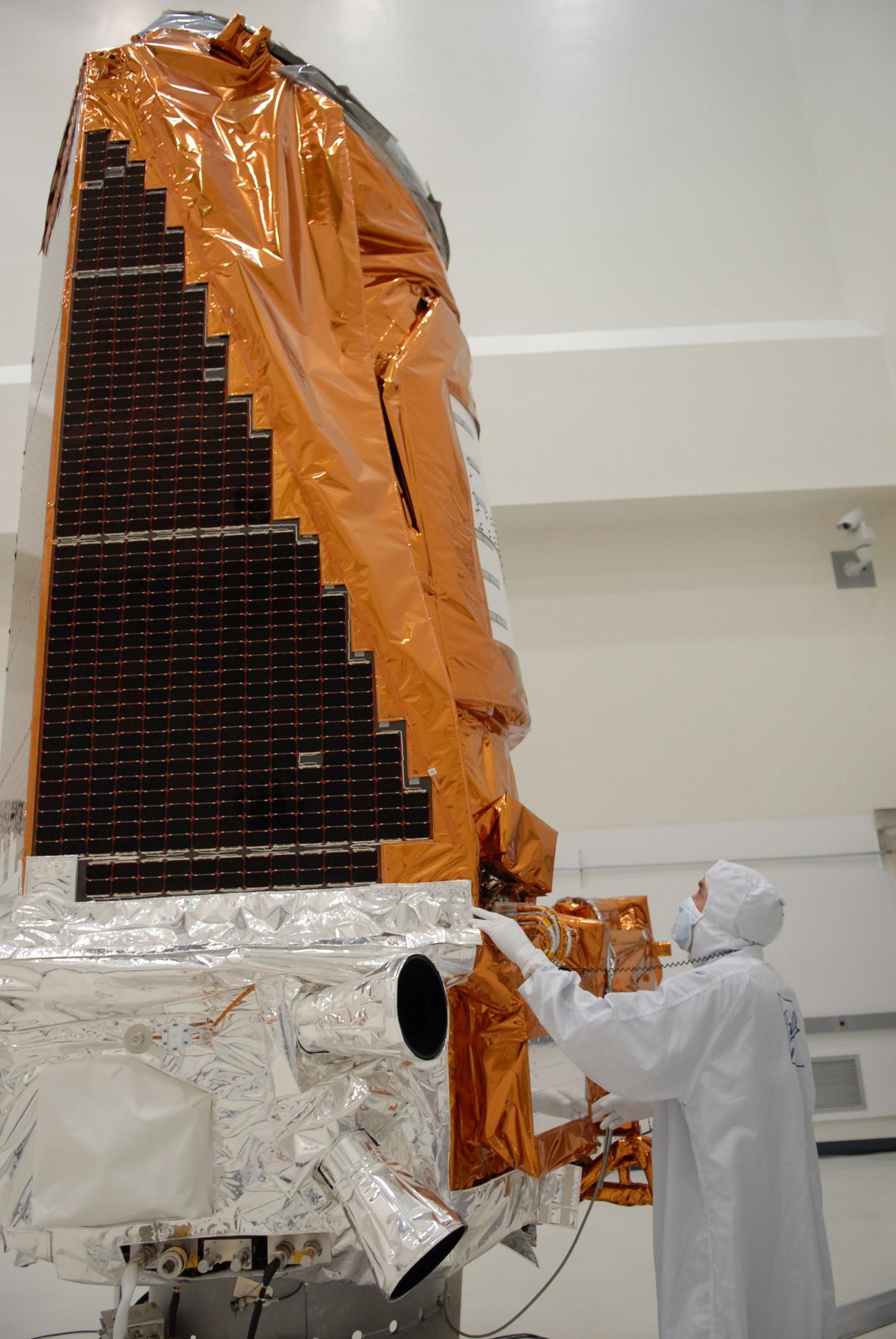 A technician in a white clean suit looks up at a large space telescope. The telescope is bronze and silver with black solar arrays.