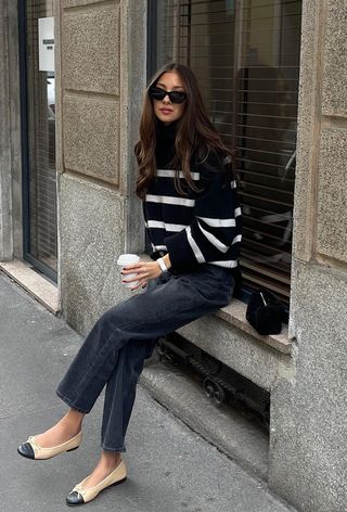 Black turtleneck outfit is shown in a photo of a French woman sitting on a window seal wearing a black striped turtleneck with black jeans, two-tone ballet flats, and a black handbag