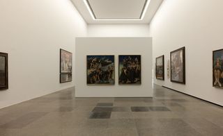 The interior of an art gallerry featuring white walls with art, white ceiling and grey square design flooring. Center white panel with arts hanging