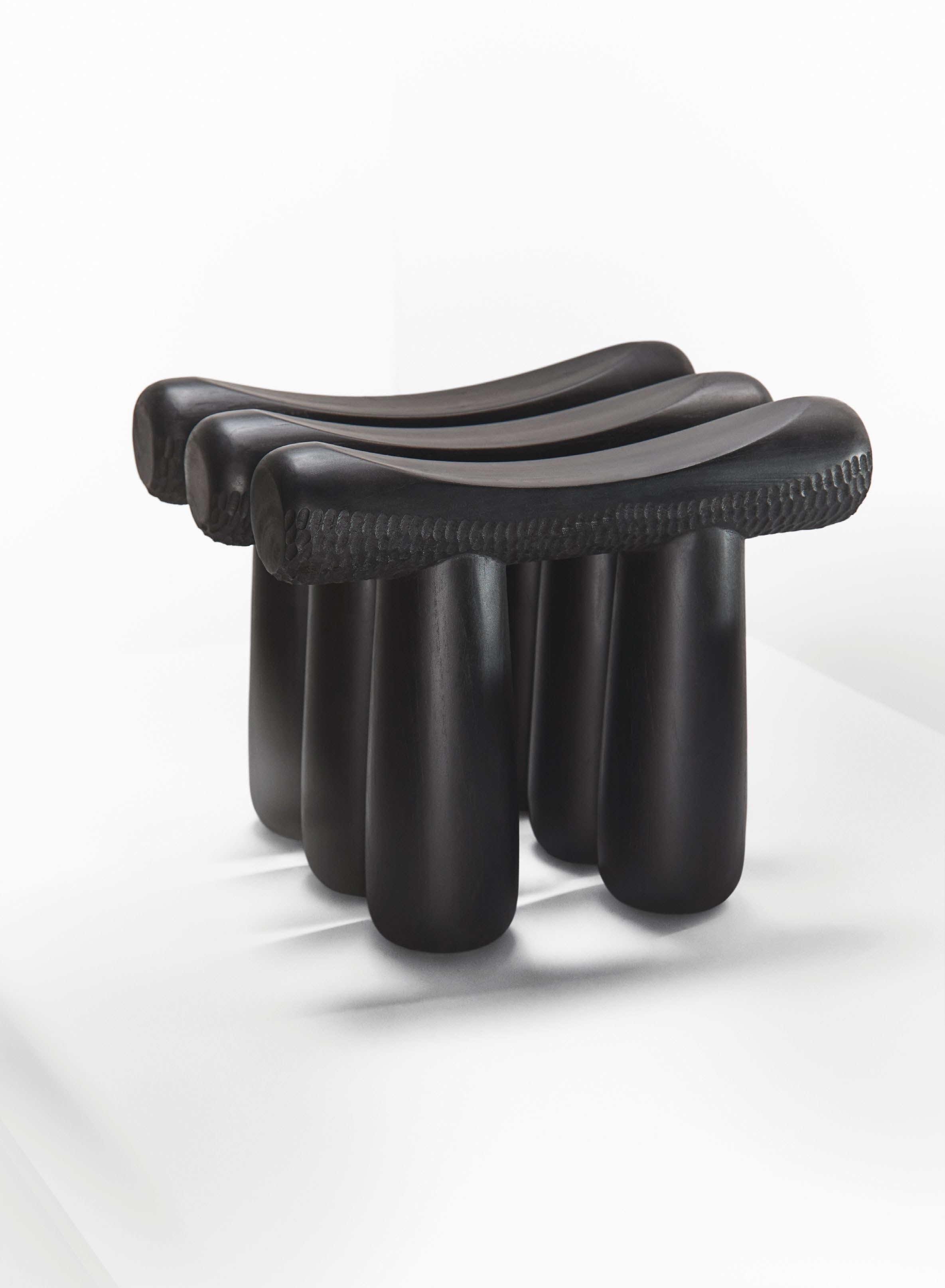 A sculptural black stool designed by Olorunfemi Adewuyi, featuring a unique form with curved surfaces and cylindrical legs, showcased in the context of the article 'MATERIAL ALCHEMISTS'.