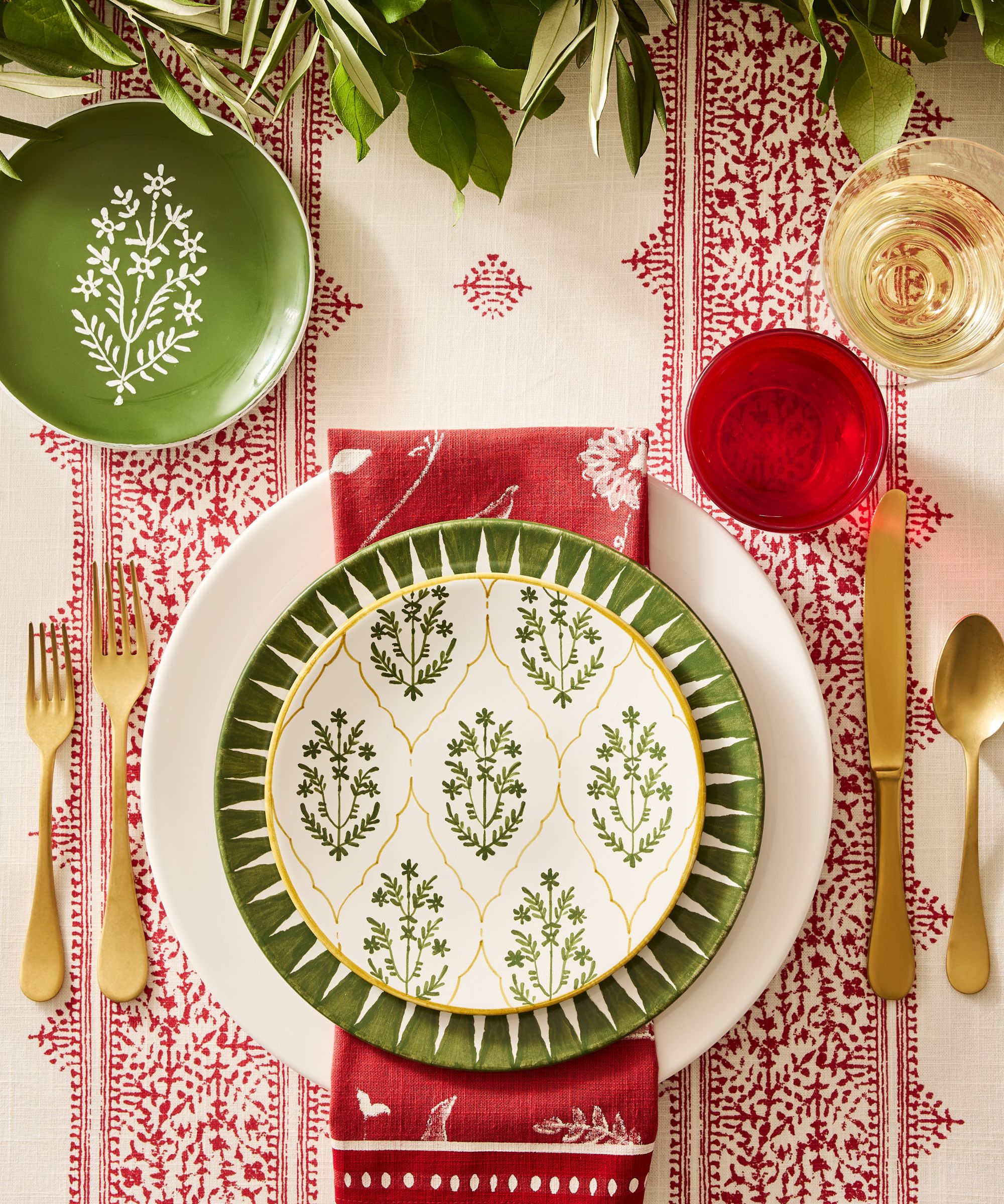 Green and red table setting with patterned plates, gold flatware and greenery