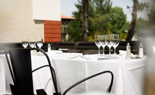 Daytime, outside image of Hotel Ville d'Hiver, Arachon dining area, black chair, table with white cloth, rows of empty glasses, cutlery, white and red brick wall, blurred trees and surrounding area, blue sky