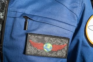 a mission patch of earth with wings, on a gray background, attached to a blue flightsuit