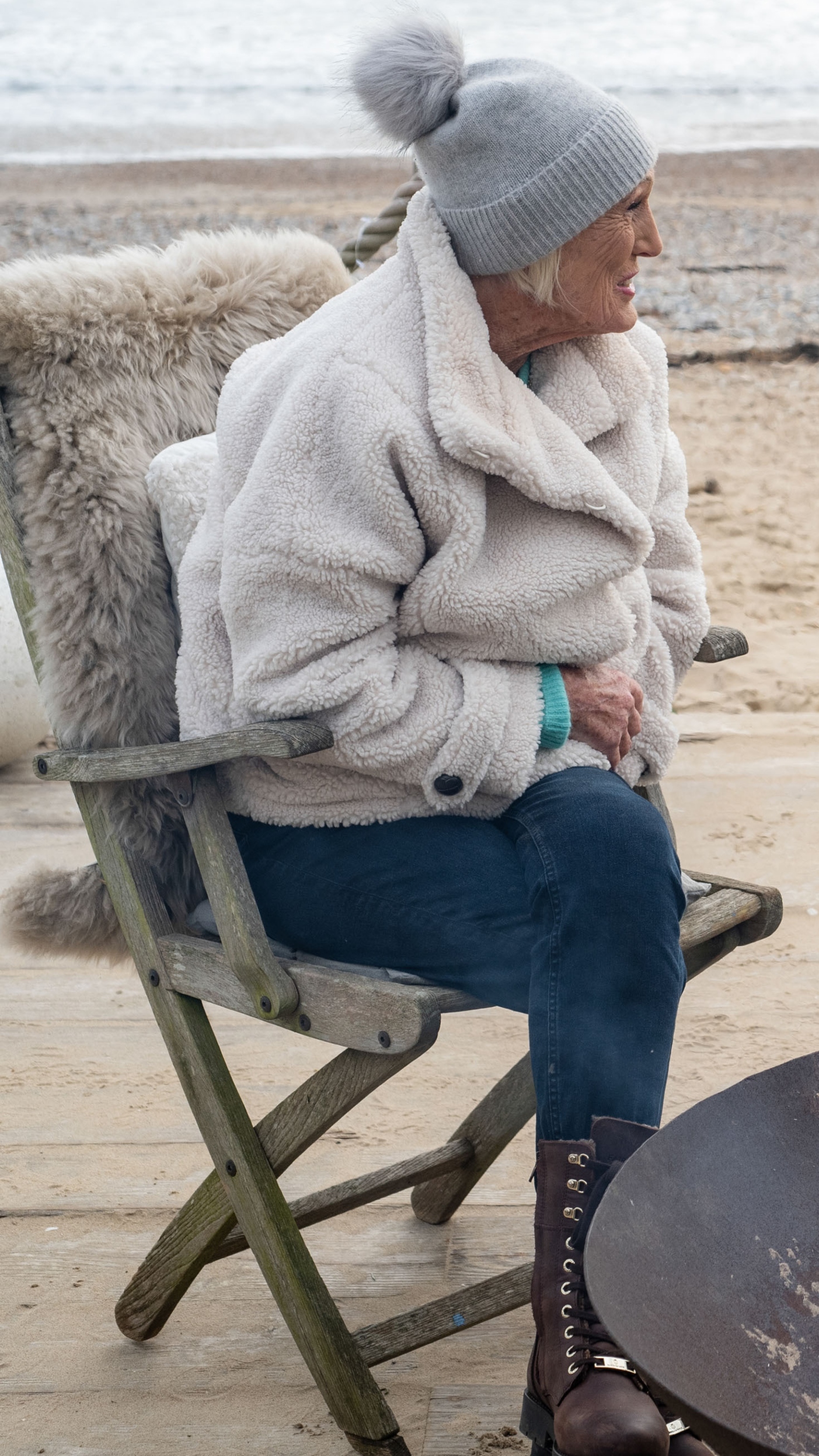 Mary Berry sits by a fire wearing jeans, shearling jacket and a bobble hat