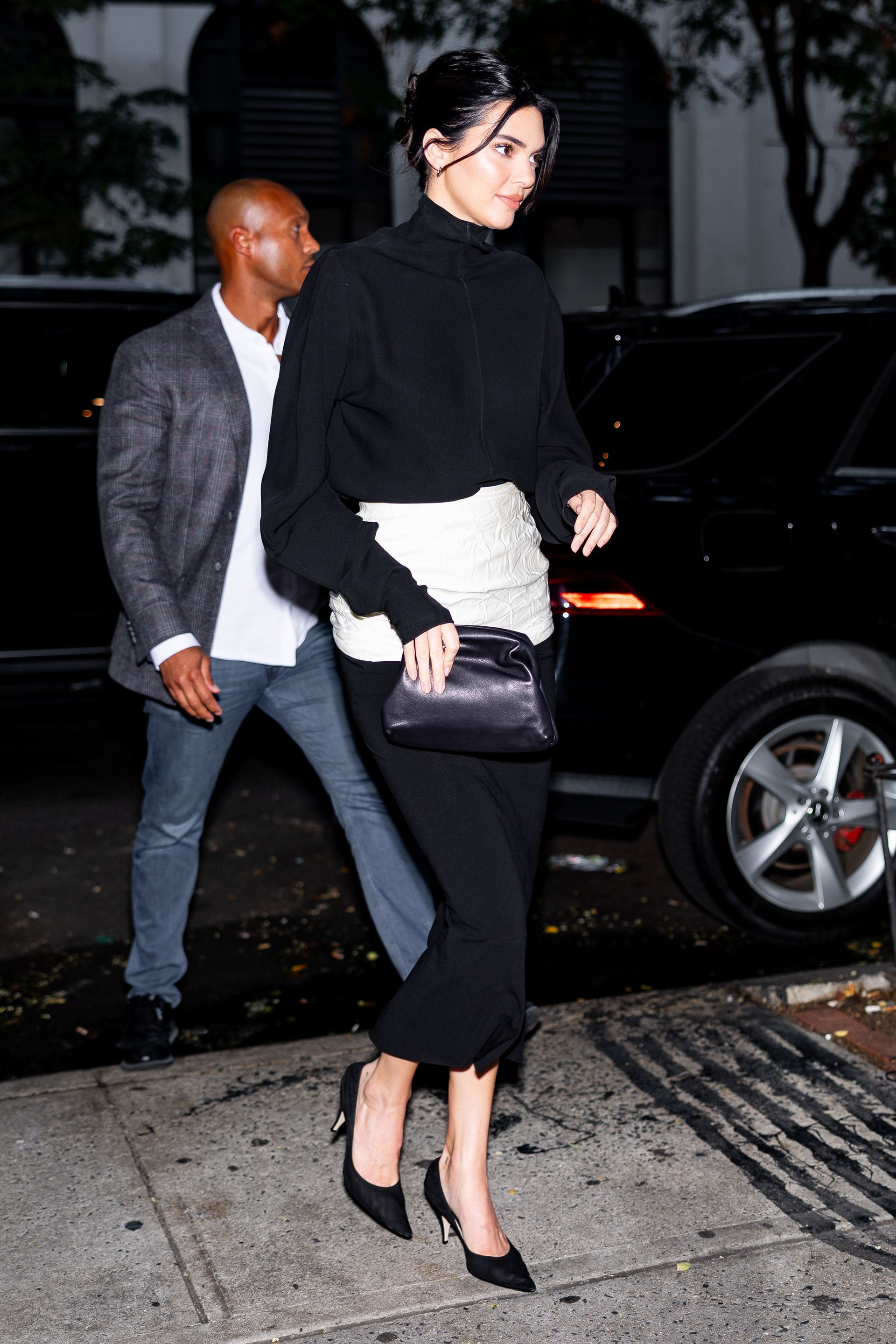 Kendall Jenner walks outside wearing a black knit, black midi skirt and mid-heel pumps.