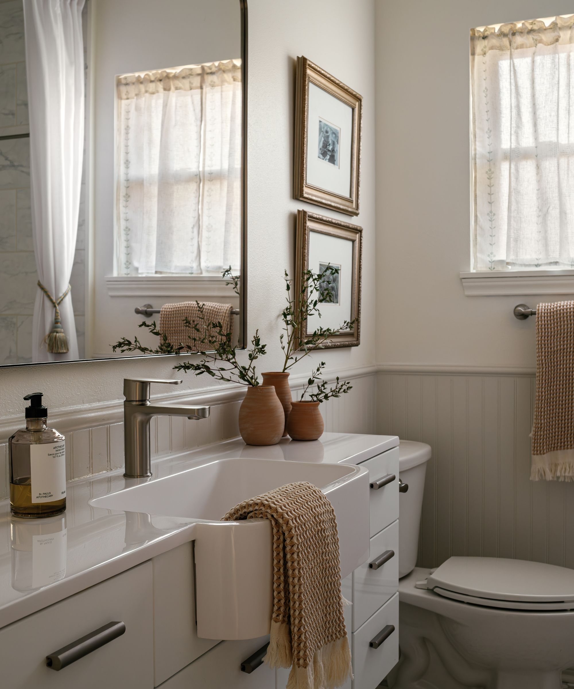 A small bathroom with light gray panelling, white walls above, and cozy rustic decor, including a cafe curtain and terracotta plant pots.