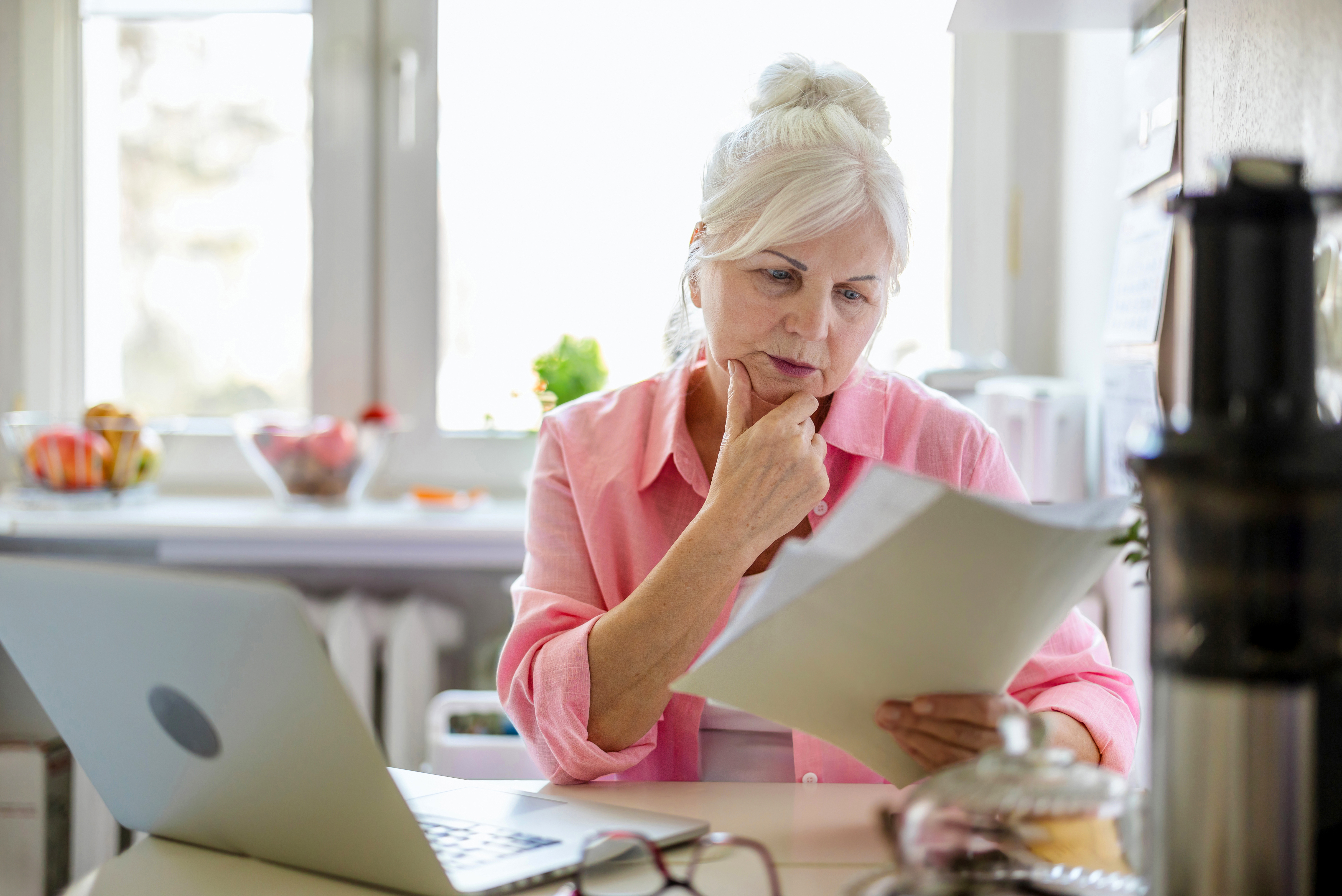 Woman looks at inheritance tax documents on paper as she sits beside laptop at kitchen table.