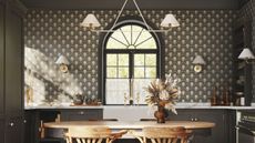 Black kitchen ideas are so chic. Here is a black kitchen with art deco wallpaper, a white hanging light with two lampshades, black cabinets and a gold faucet above a white sink, and a curved light wooden dining table with flowers on and two chairs