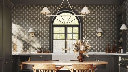 A black kitchen with art deco wallpaper, a white hanging light with two lampshades, black cabinets and a gold faucet above a white sink, and a curved light wooden dining table with flowers on and two chairs