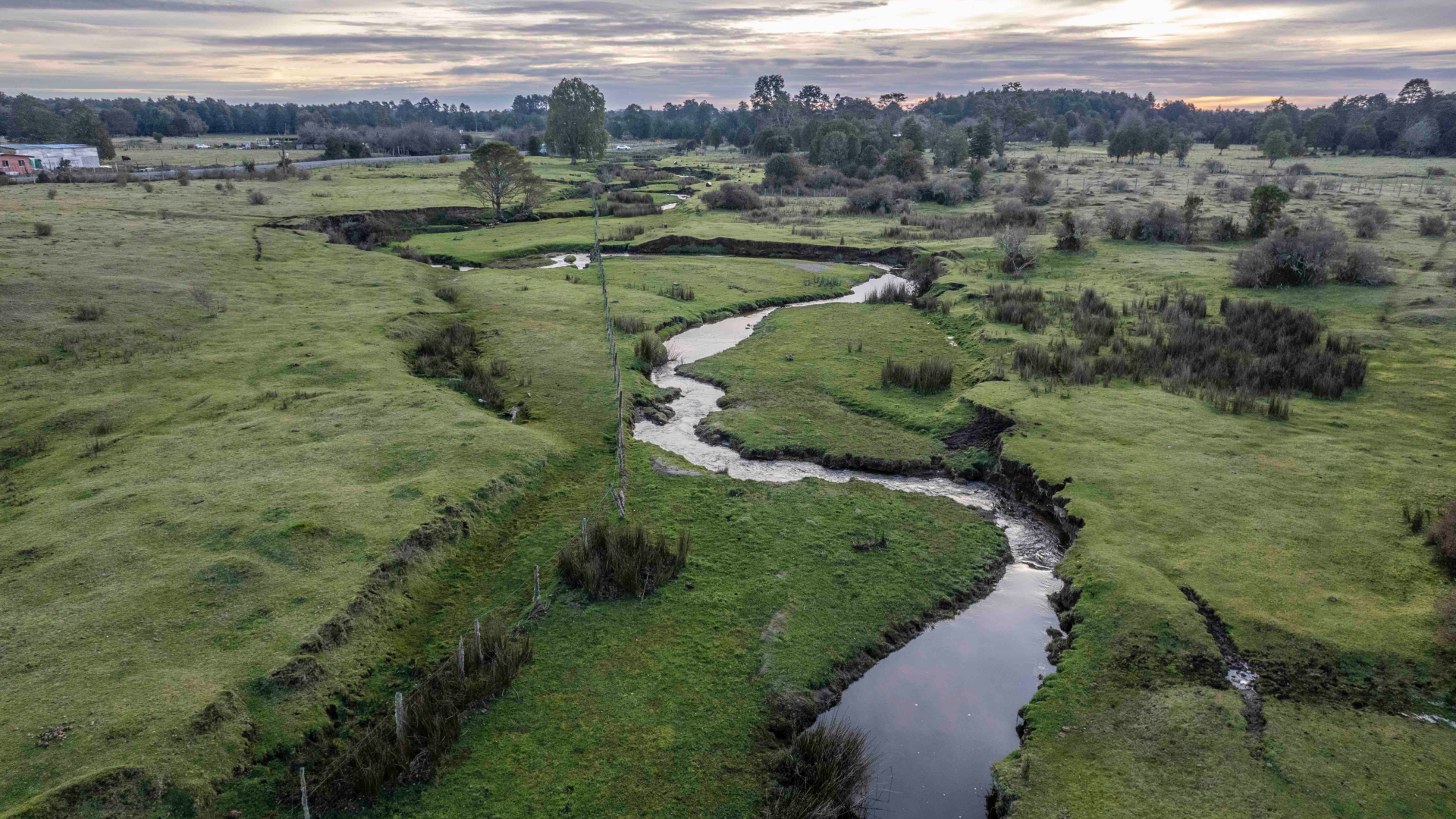 a green landscape cut through the middle by a meandering stream with a cloud-strewn sunset in the background