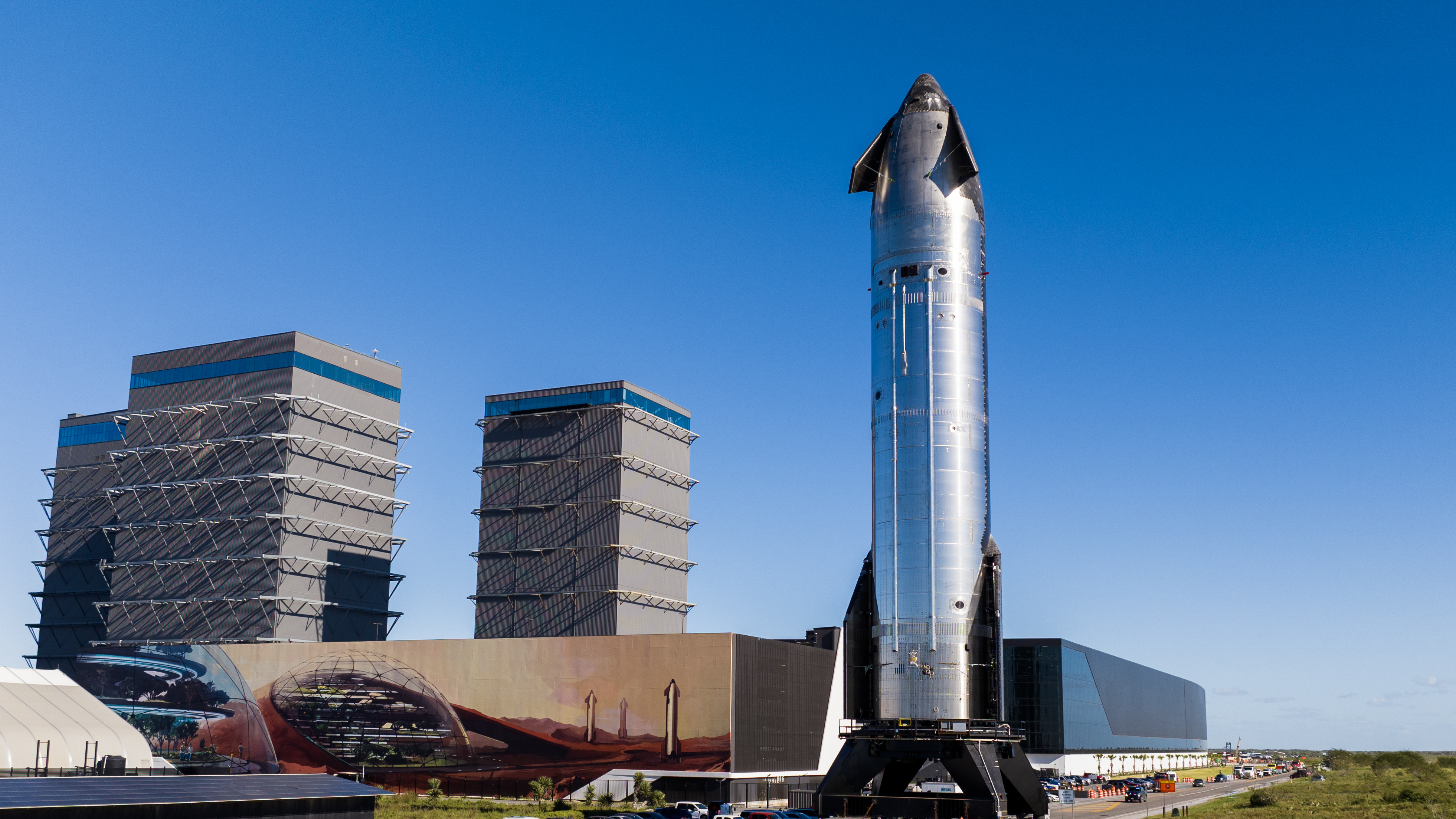 a chrome spaceship stands on in front of three tall rectangular buildings.