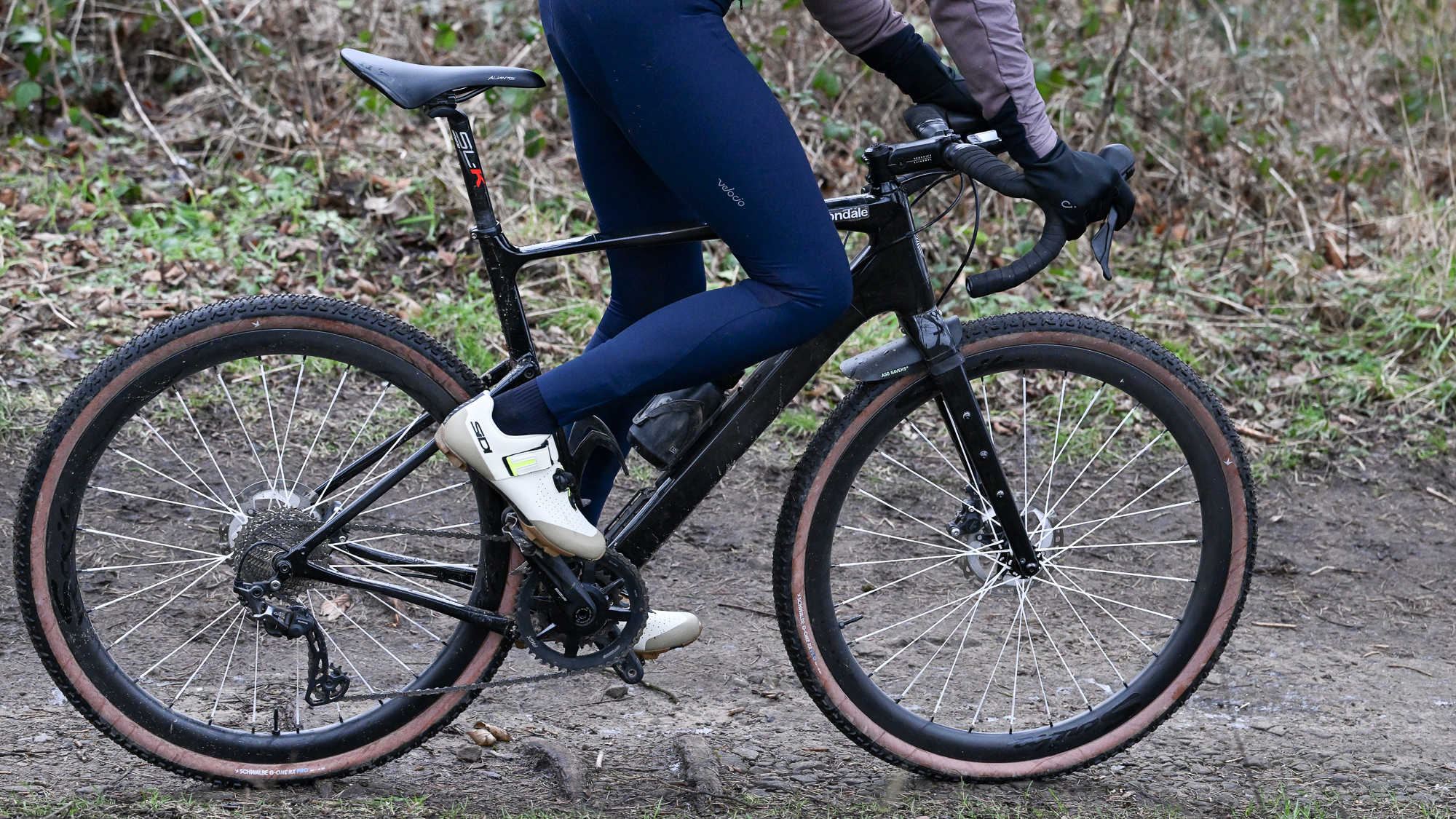 Side view of a man's lower body riding a gravel bike on a dirt track wearing blue tight sand beige shoes