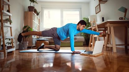 man wearing a blue long sleeved top and grey shorts performing a pilates core move like a bird dog in a home setting on an exercise mat on a wooden floor in front of a laptop. 