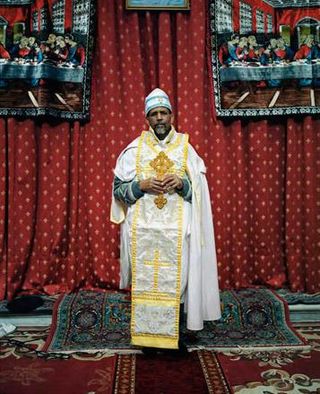 Bearded priest in white and gold robes pictured against red and gold curtains and standing on a red Persian-like rug.