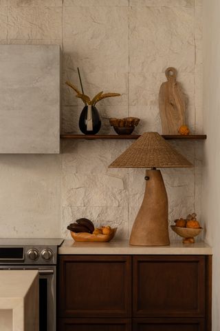 A wood and rattan fantastically shaped lamp sits atop a kitchen surface in cream marble and wood surrounded by fruit baskets filled with oranges.