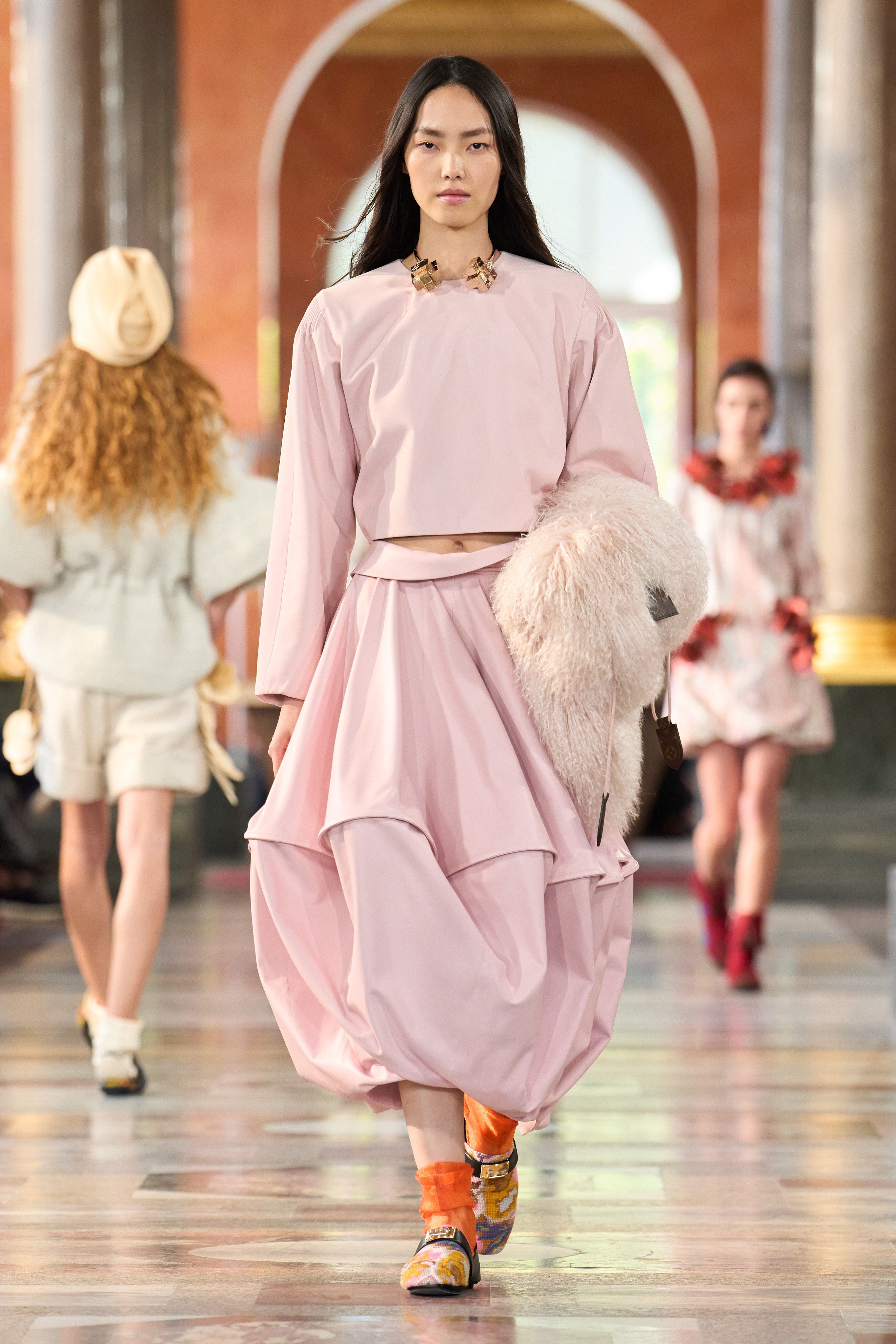 A Louis Vuitton model wearing a pink crewneck long-sleeve blouse with a matching light pink balloon skirt, a chunky gold collar, a shaggy pink shearling sole, and bright orange sandals.