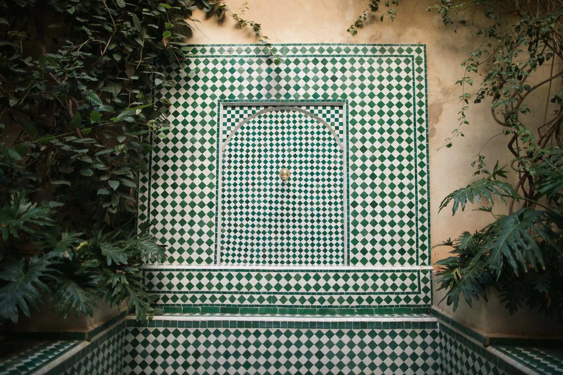 A tiled fountain with seagreen and white tiles sits in an inner courtyard filled with verdant foliage.