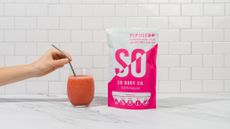 Packet of So Body Co. next to red smoothie in small glass with a hand holding a straw
