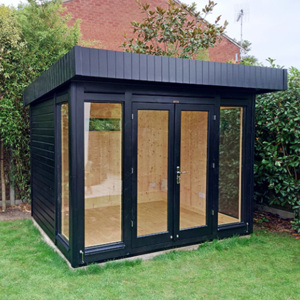 A black timber-clad flat-roof garden room with full-height glazed double doors revealing an empty pine-lined interior, set on a lawn against a hedge.