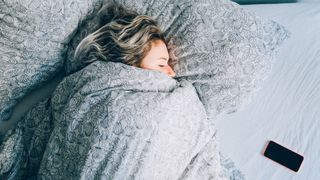 Woman snuggled up in a duvet in bed, with a phone next to her