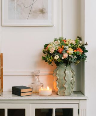 A white entryway with a glass vase filled with flowers, a candle, and a box on top of a white console table