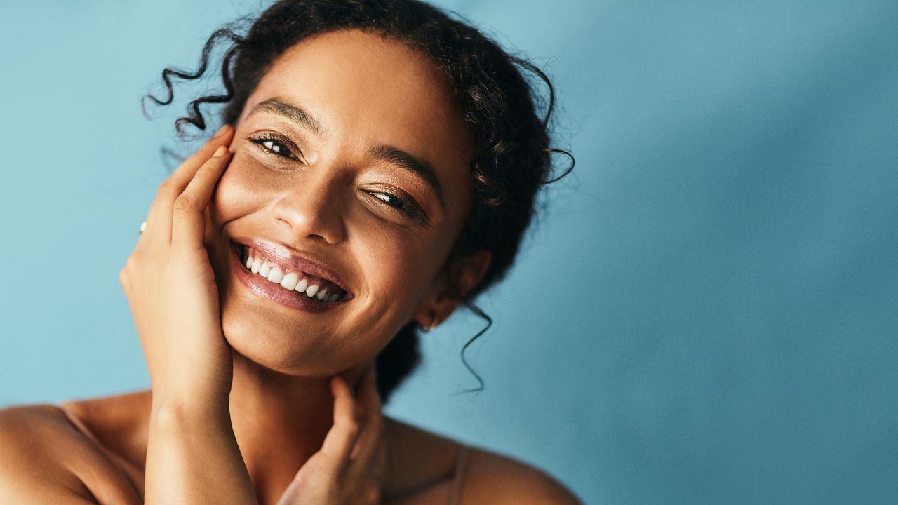 Elemis Dynamic Resurfacing Facial Wash - beautiful woman with her hand on her cheek smiling against a blue backdrop gettyimages:1181806328