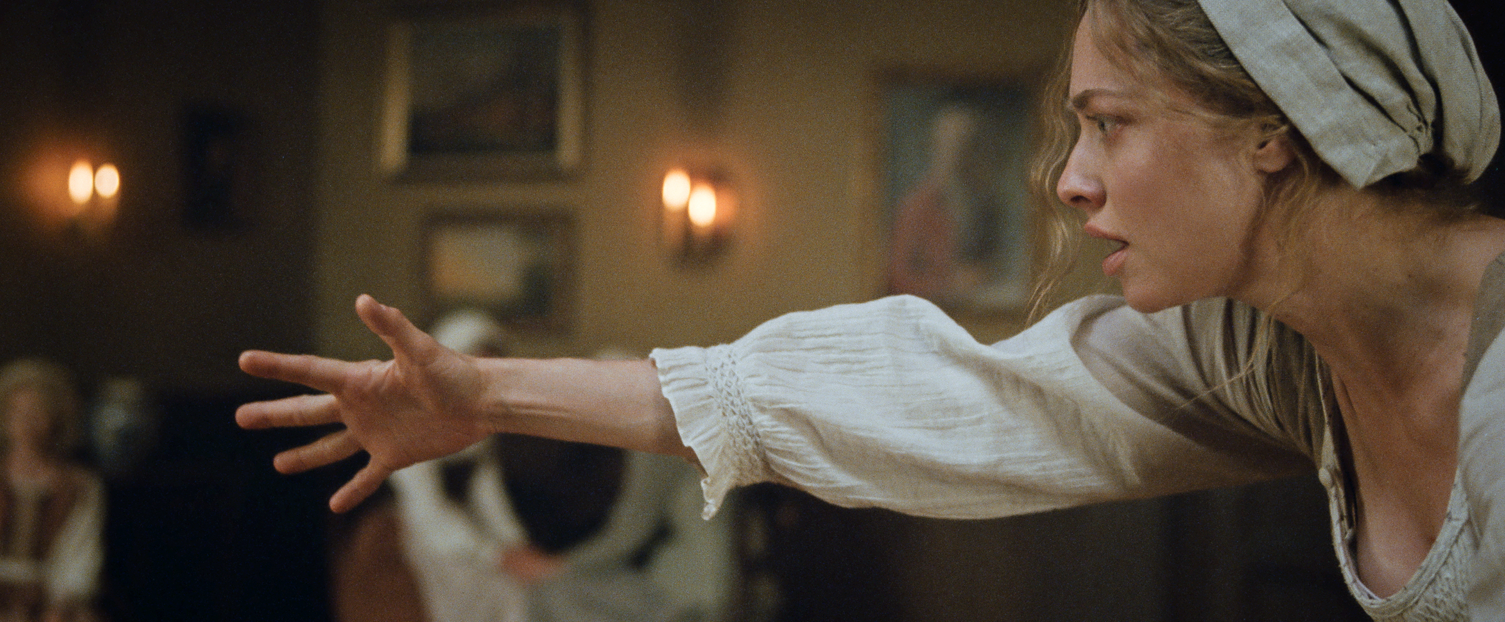 A close-up on a blond woman (Amanda Seyfried as Ann Lee) wears a bonnet and 18th-century blouse. She stands to the right and extends her hand left across the frame, as blurred figures stand in the background, in a still from in THE TESTAMENT OF ANN LEE.