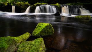 Landscape photo of a river and waterfall with green mossy rocks in the foreground
