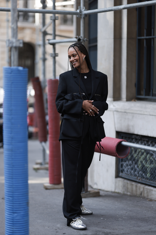 A guest is seen wearing black sunglasses, a black shiny suit with big attached bags and zippers on the blazer combined with black Botter shirt with white B on the top, lots of silver rings and silver earrings and silver shiny Adidas x Wales Bonner sneaker outside Botter show during the Menswear Spring/Summer 2024 as part of Paris Fashion Week on June 21, 2023 in Paris, France.