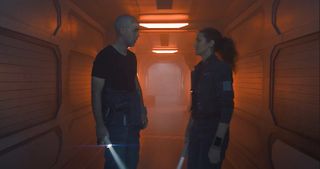 A man and woman face off in a spaceship corridor bathed in orange-red light