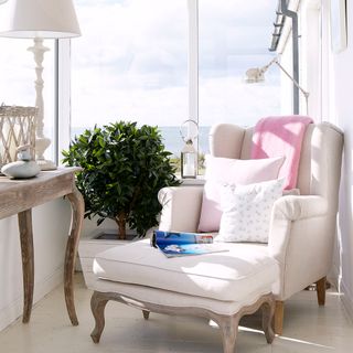 an elegant white armchair with matching foostool with wooden legs tucked in the corner of a small white conservatory, with a matching wooden side table and indoor plant, by large windows overlooking the ocean