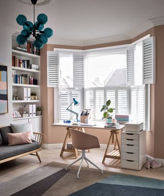 Light-filled home office set-up in living space with trestle desk in front of bay windows, dressed with white shutters.